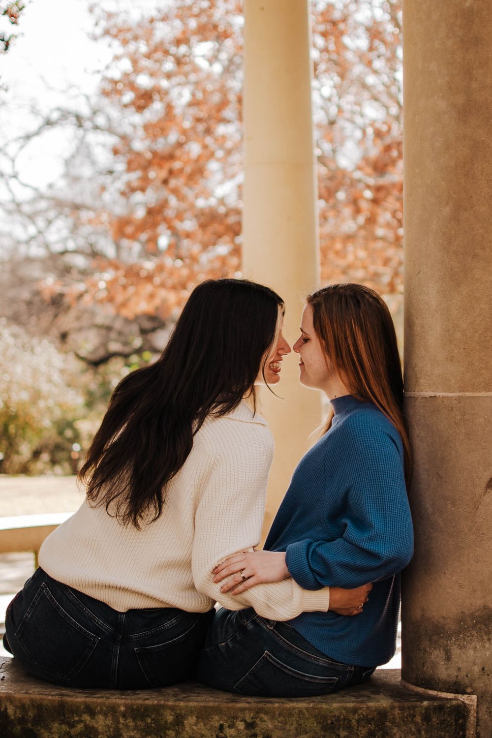 Two women in cozy sweaters sit close together, touching noses romantically, surrounded by autumn foliage and stone columns.
