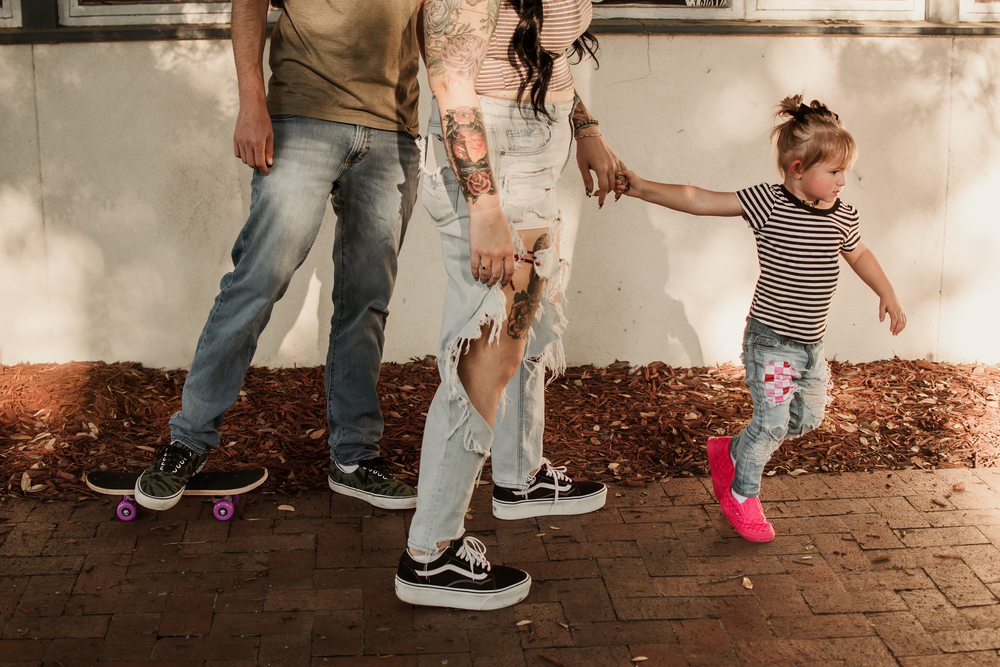 Family with tattooed adults and young girl in striped outfit walking together, wearing Vans sneakers on brick pathway.