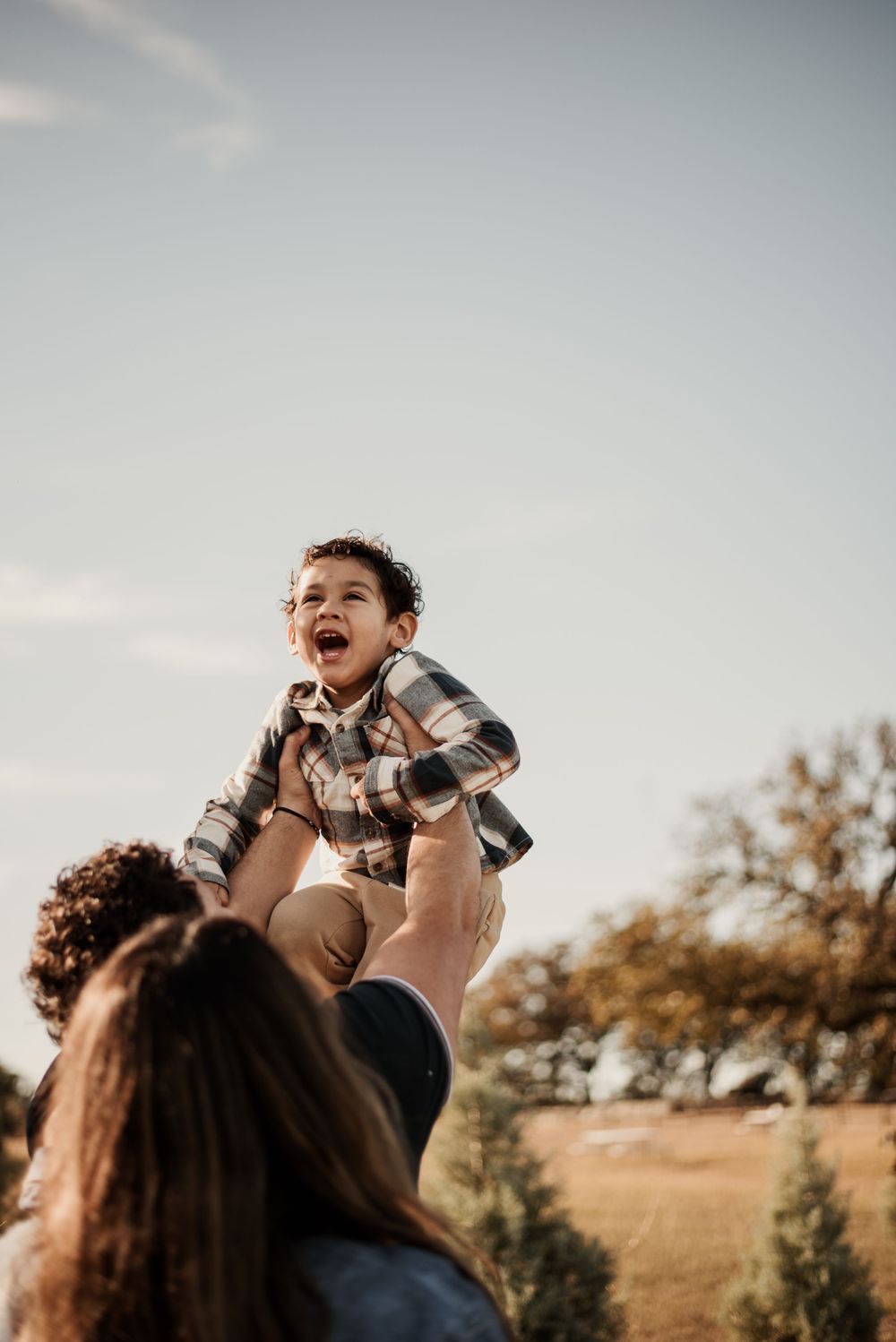 A joyful toddler in a plaid shirt is lifted high in the air by a parent outdoors on a sunny day.