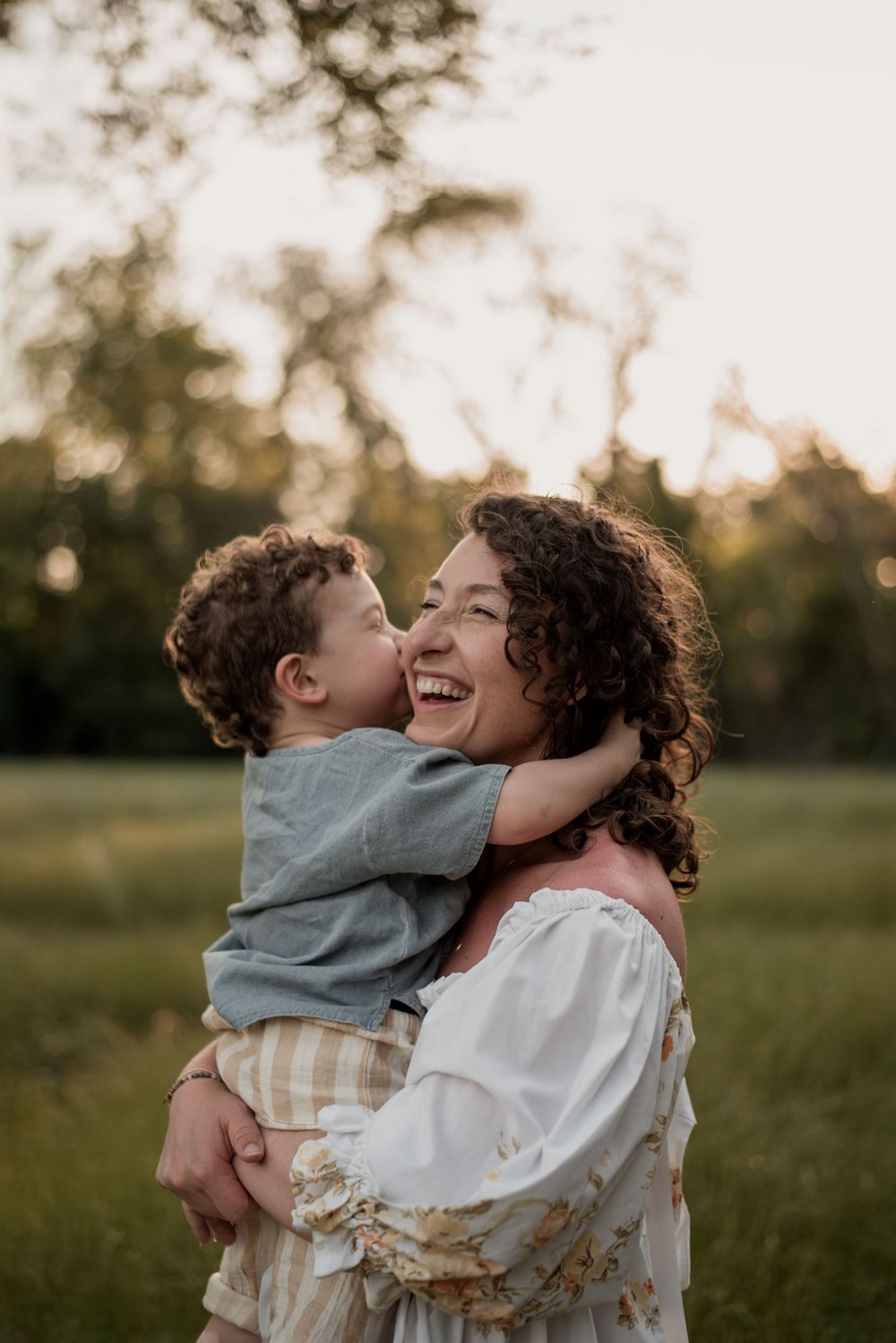 A joyful mother laughs as her curly-haired toddler gives her a kiss outdoors in a sunlit green field.