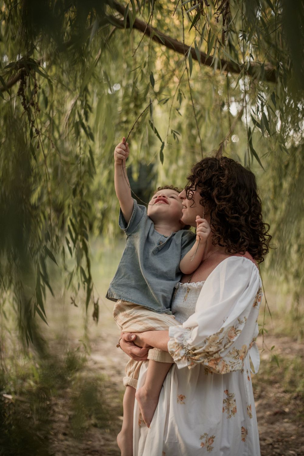 A curly-haired mother holds her young son under a weeping willow tree as he reaches up to touch a branch.