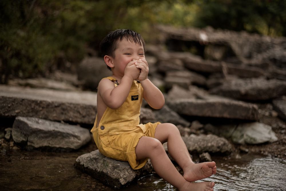 Young boy in yellow romper sits barefoot on rocks by a stream, hands covering his mouth.