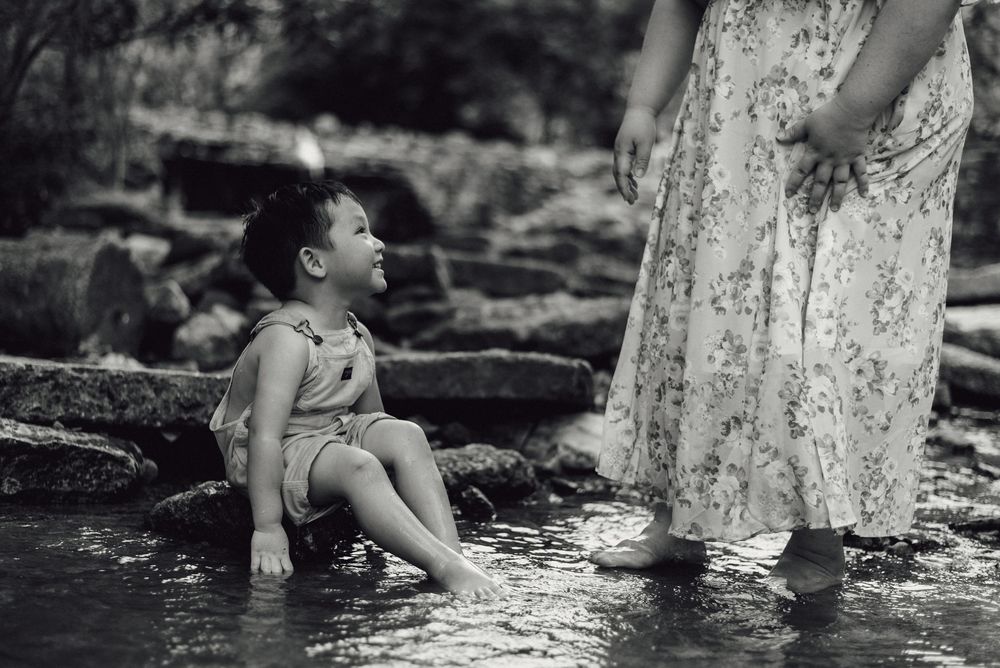 Black and white photo of a young child sitting in a shallow stream, looking up at a woman in a floral dress.