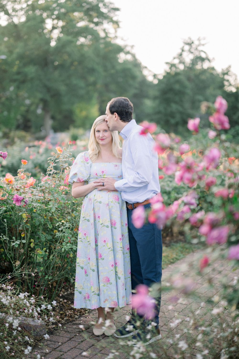 A couple shares a romantic moment in a garden filled with blooming pink and orange flowers on a stone pathway.