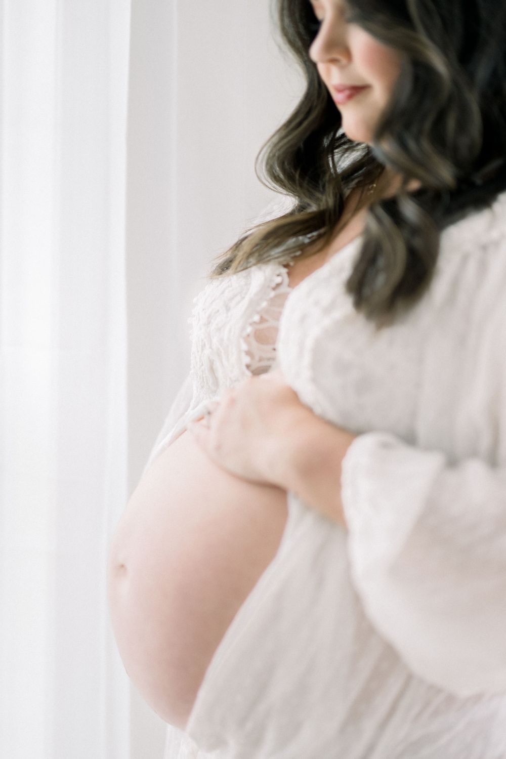 Ethereal maternity photo showing a pregnant belly in soft white lace against a bright window backdrop.