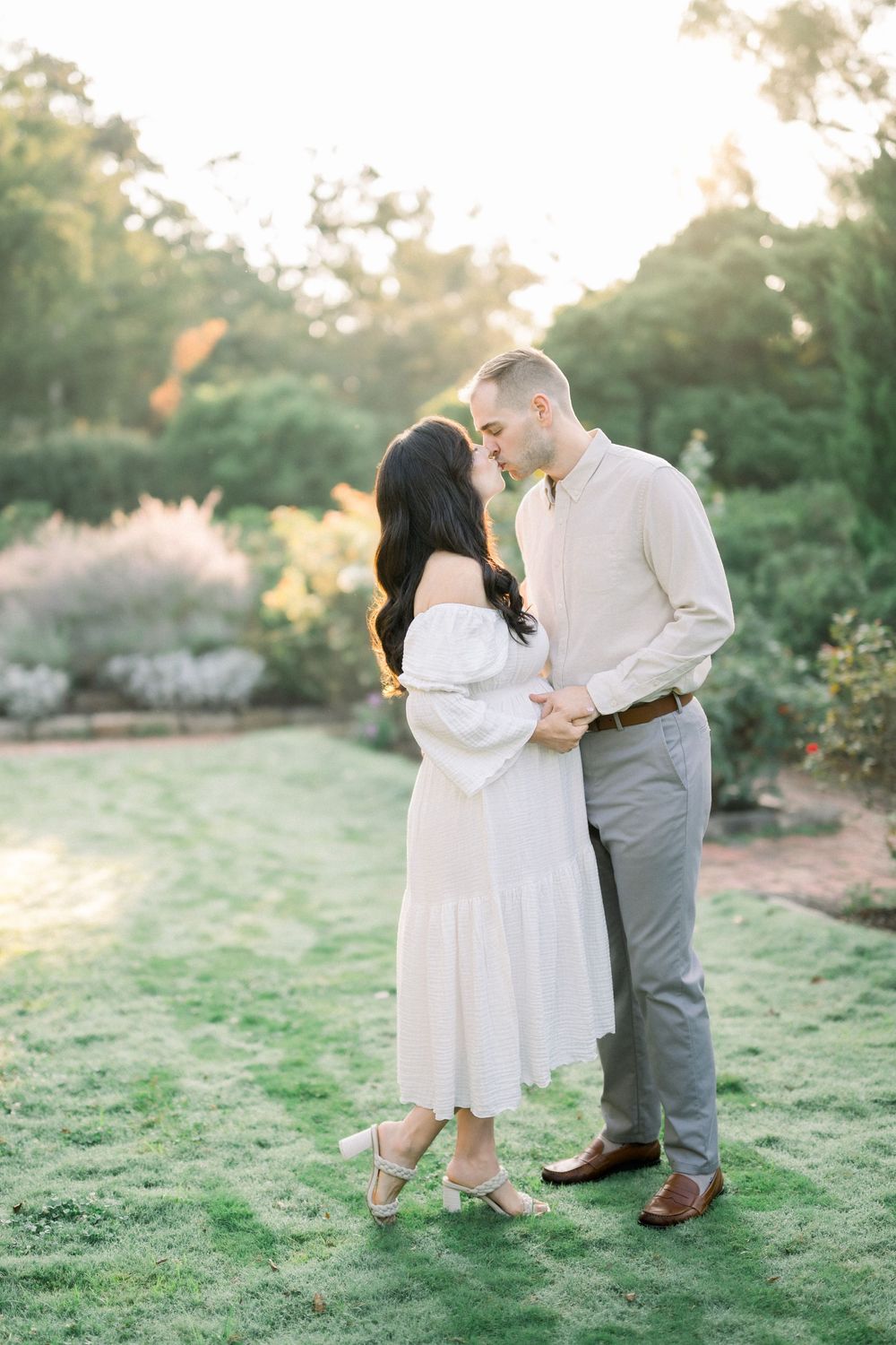 A romantic couple dances together on a grassy hill during golden hour sunset in matching white and gray outfits.