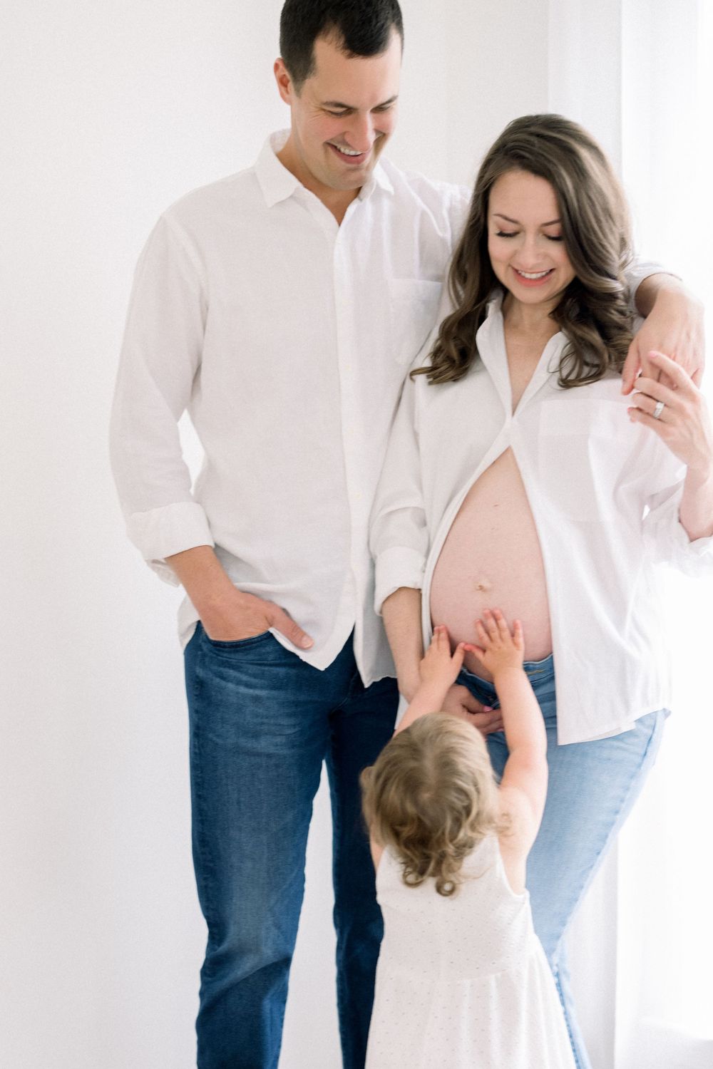 A family in white shirts and jeans laughs together against a bright white background in a lifestyle photo session.