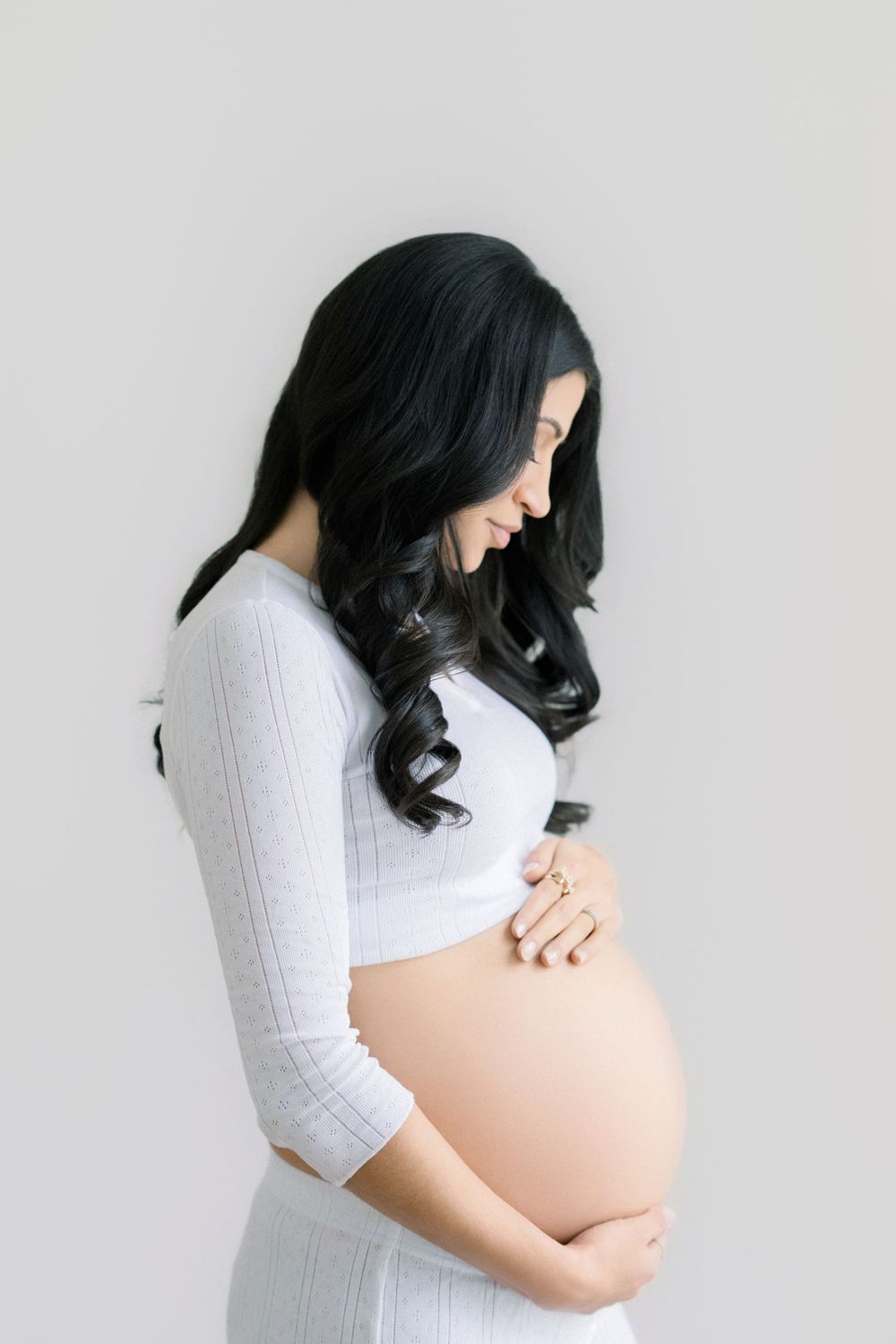 Smiling pregnant woman with dark hair cradling baby bump, wearing white ribbed crop top, studio maternity photo.