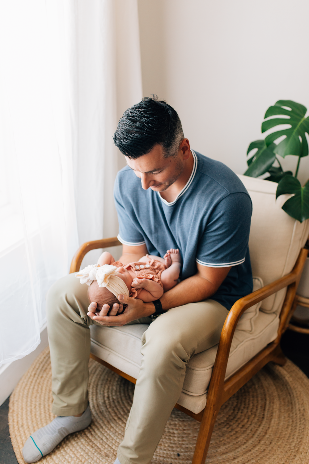 Caring parent tenderly holds newborn baby while sitting in wooden armchair near window with monstera plant.