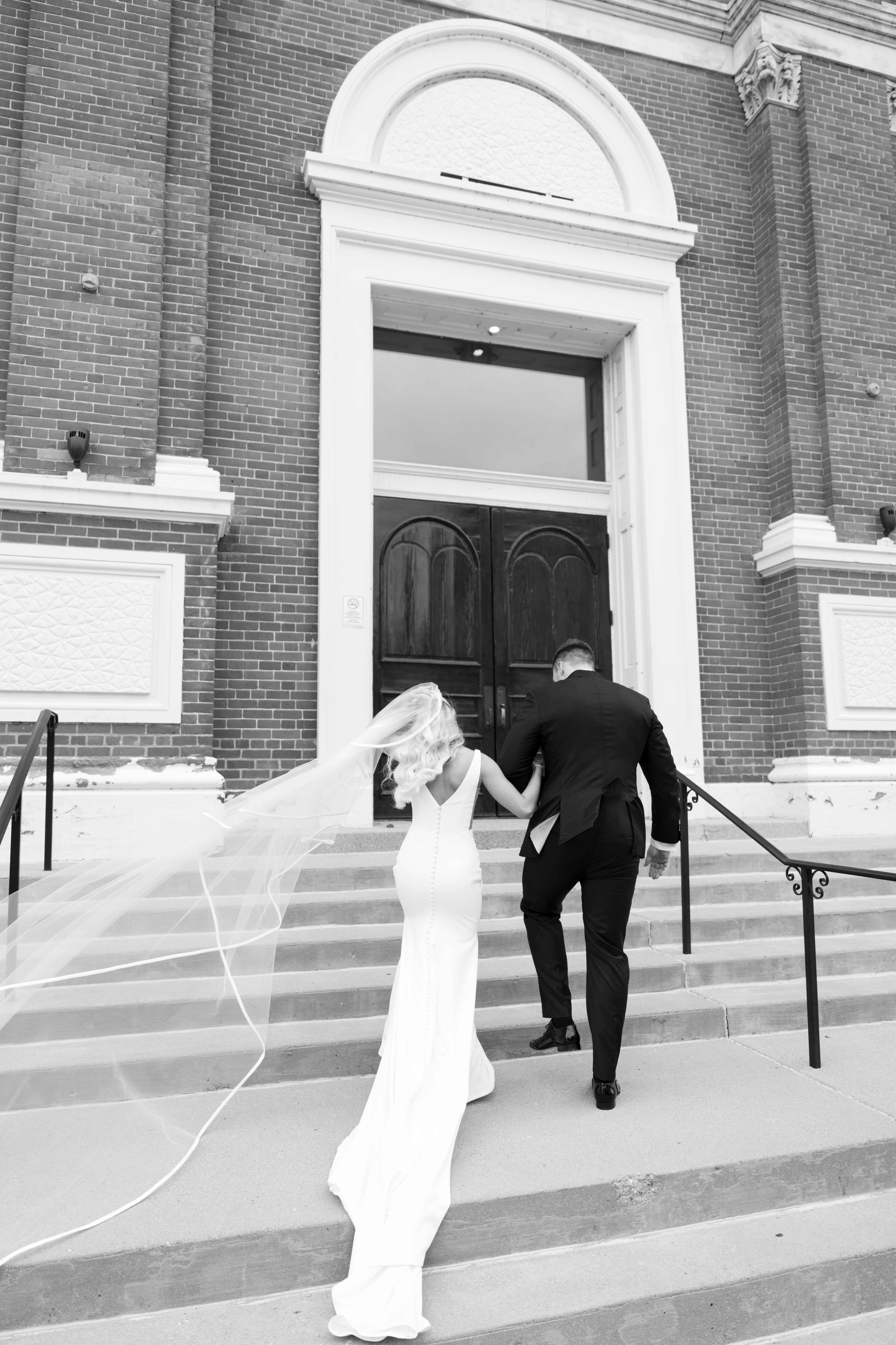 Newlywed couple climbing church steps together while bride's veil flows in the wind in black and white photo.