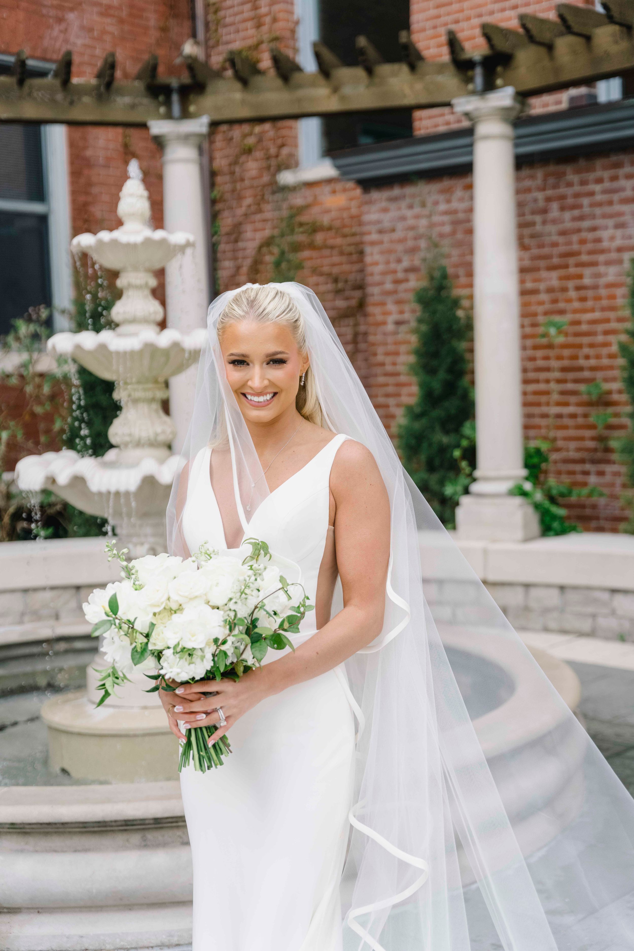 A bride in an elegant white gown holds a white floral bouquet while standing near a stone fountain at a red brick venue.