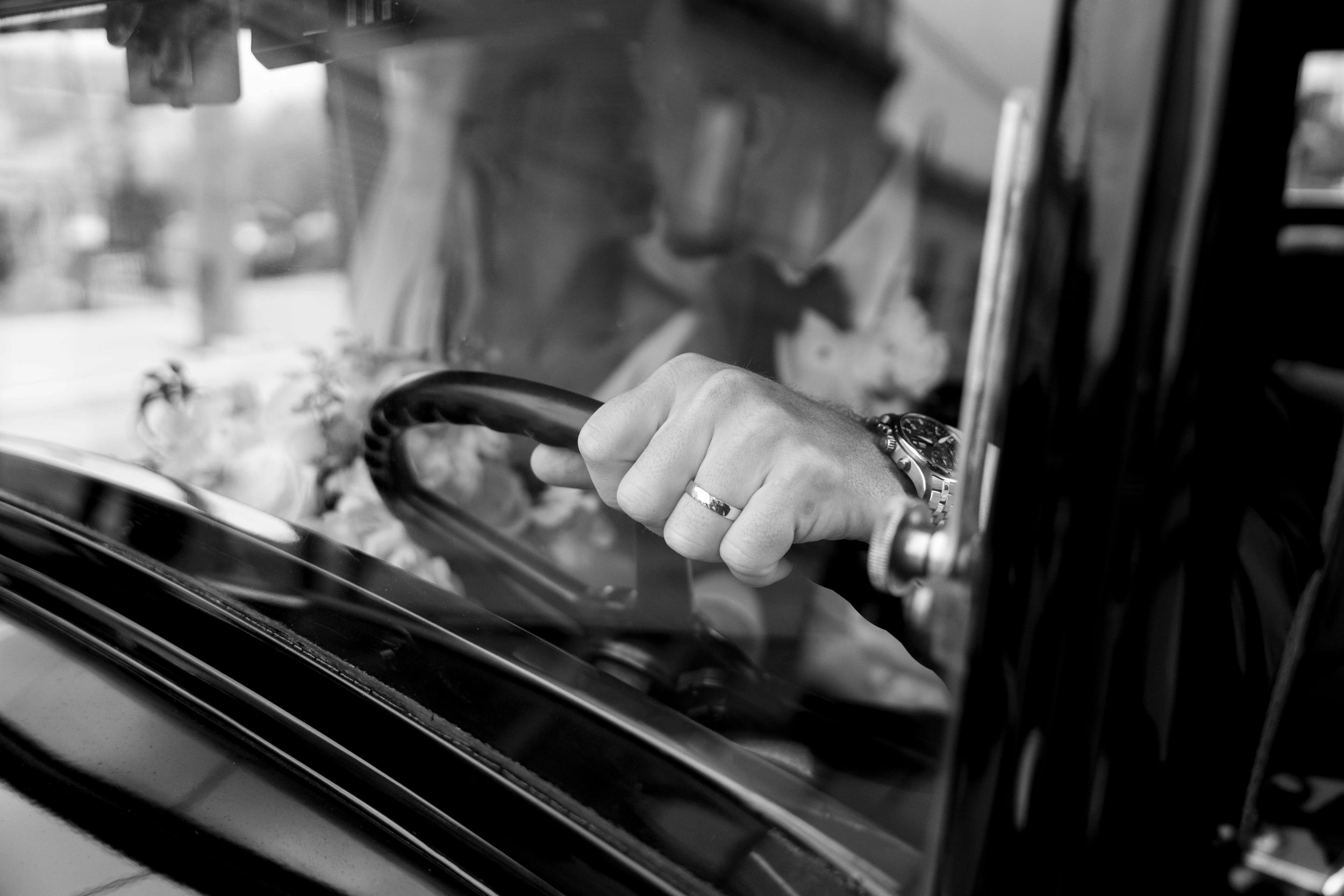 Black and white close-up of hands gripping a car steering wheel through a windshield.