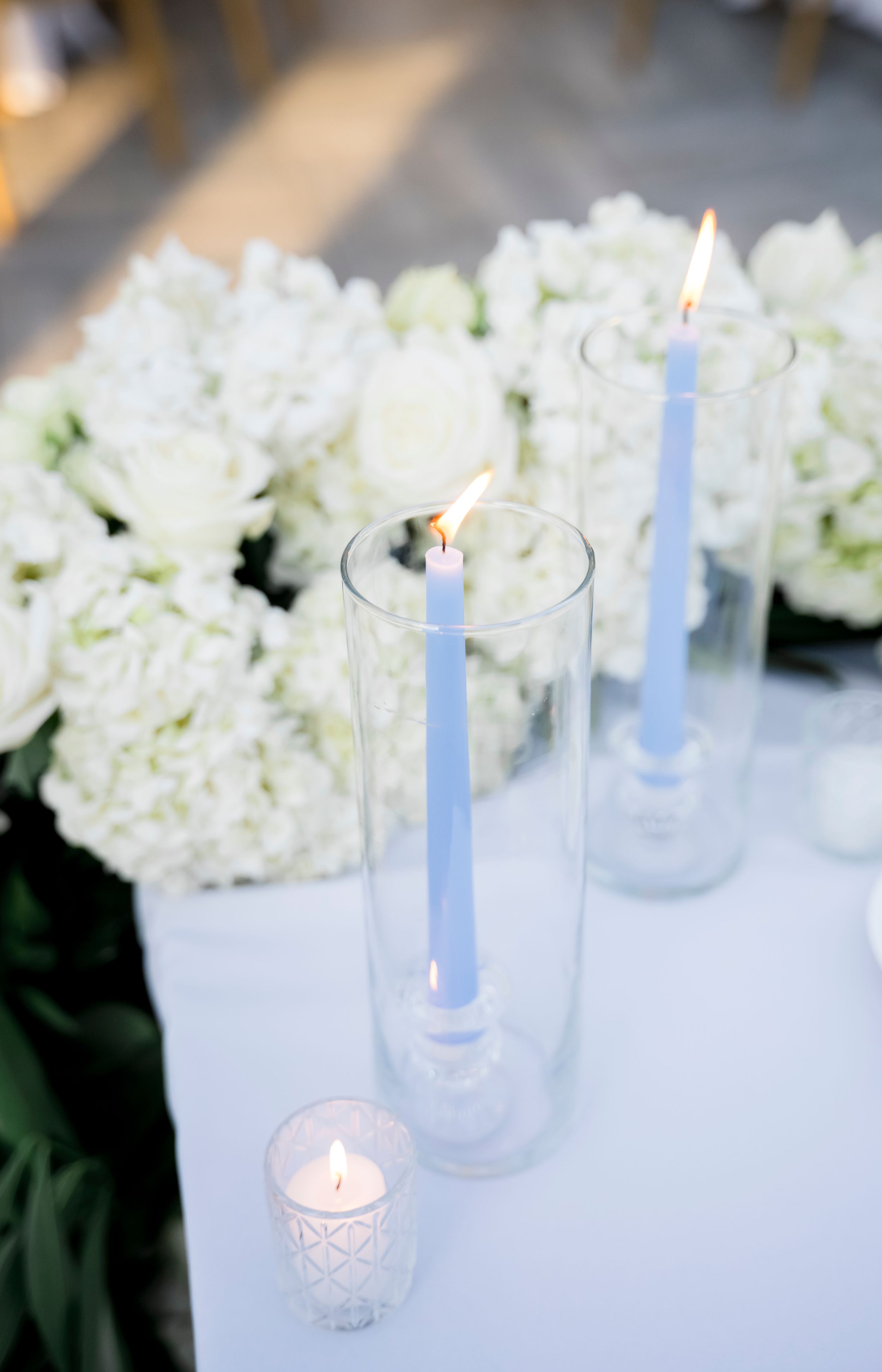 Light blue taper candles in glass holders alongside white floral arrangements create an elegant table setting.
