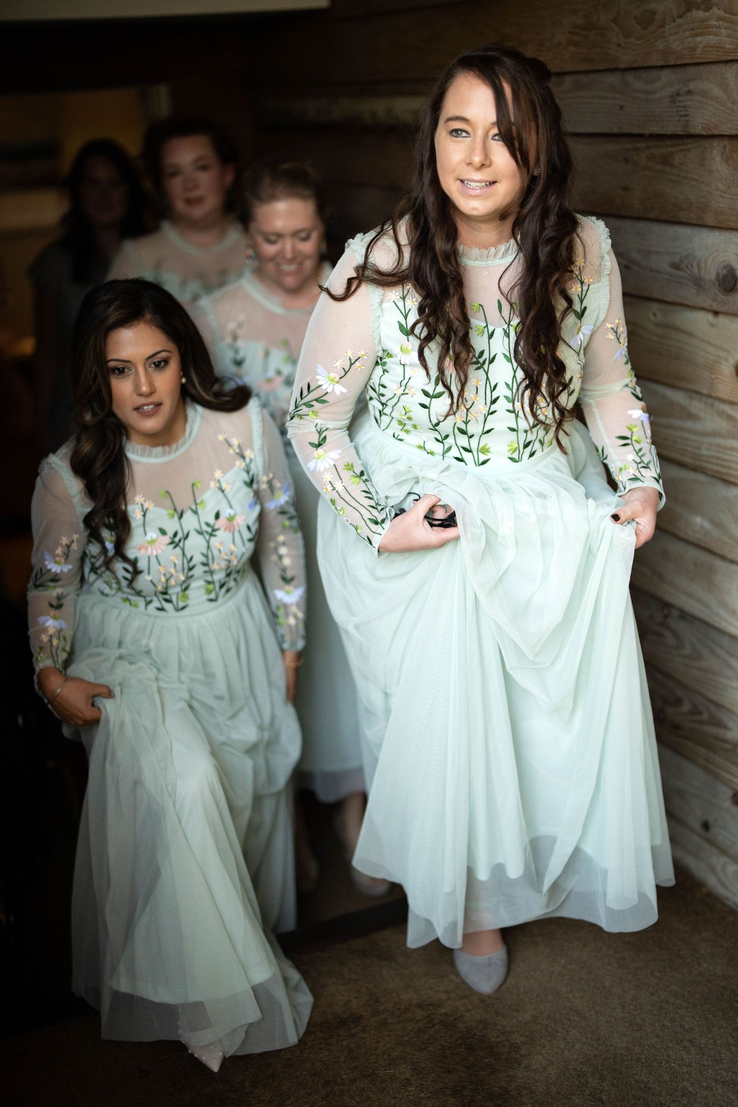 Bridesmaids dressed in elegant white flowy gowns with delicate floral embroidery walk down rustic wooden stairs.