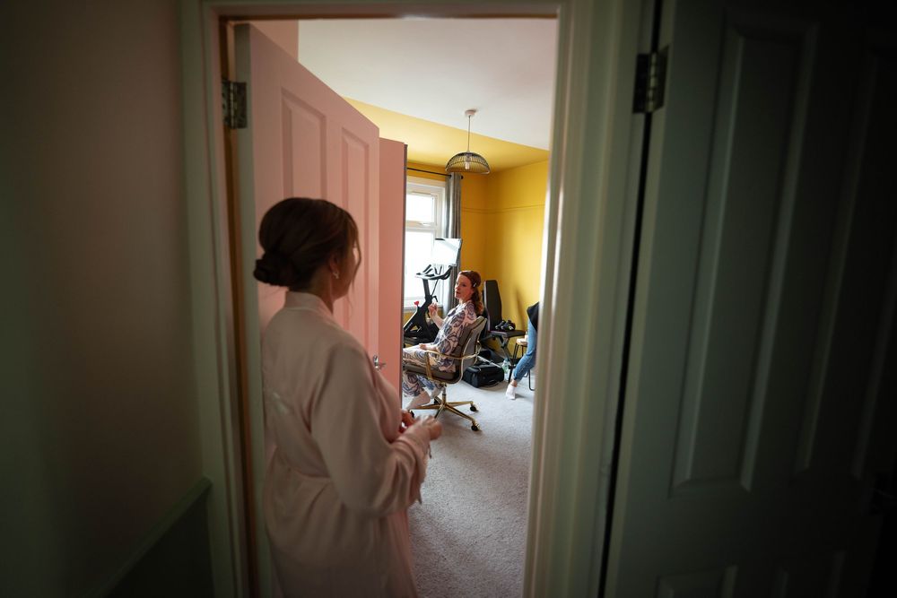 Through a doorway, someone in a bathrobe stands at the entrance of a yellow-walled room while bride has hair done