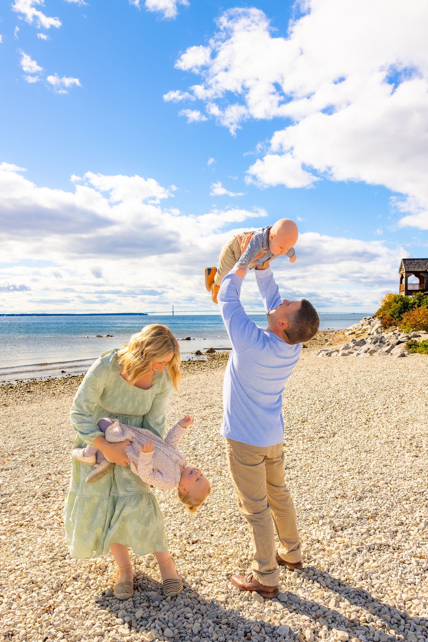 Family enjoying a sunny day at the beach, playing and lifting babies against a backdrop of blue sky and ocean.