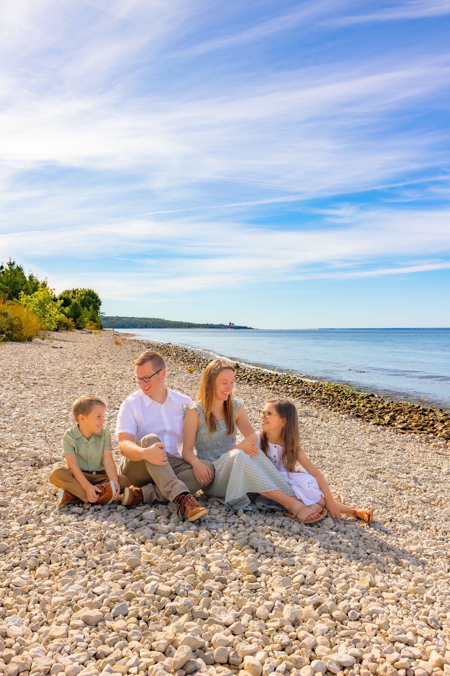 A family relaxes together on a rocky beach shoreline with blue skies and calm waters in the background.