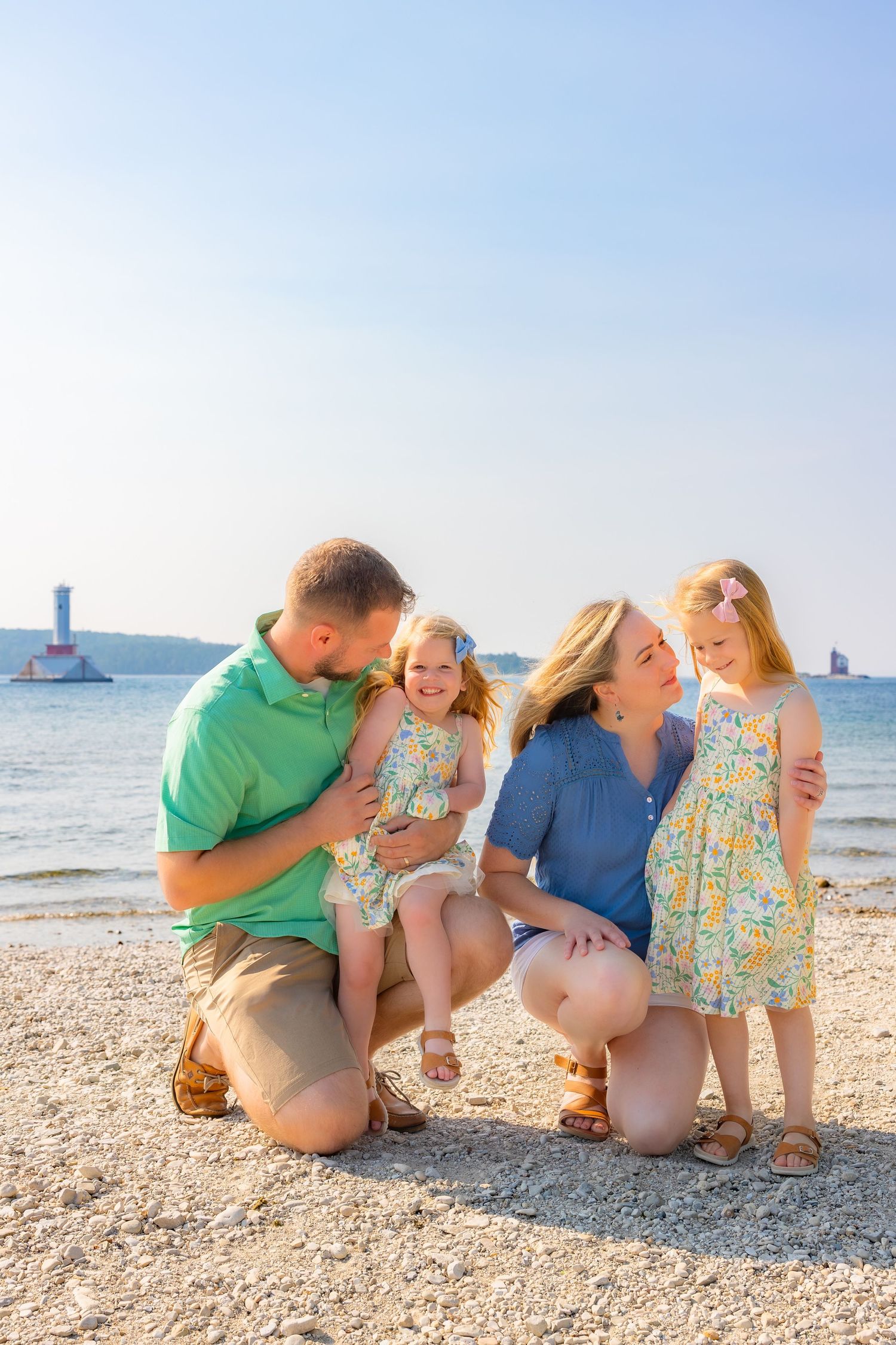 A family shares joyful moments together while kneeling on a sunny beach with ocean waves in the background.