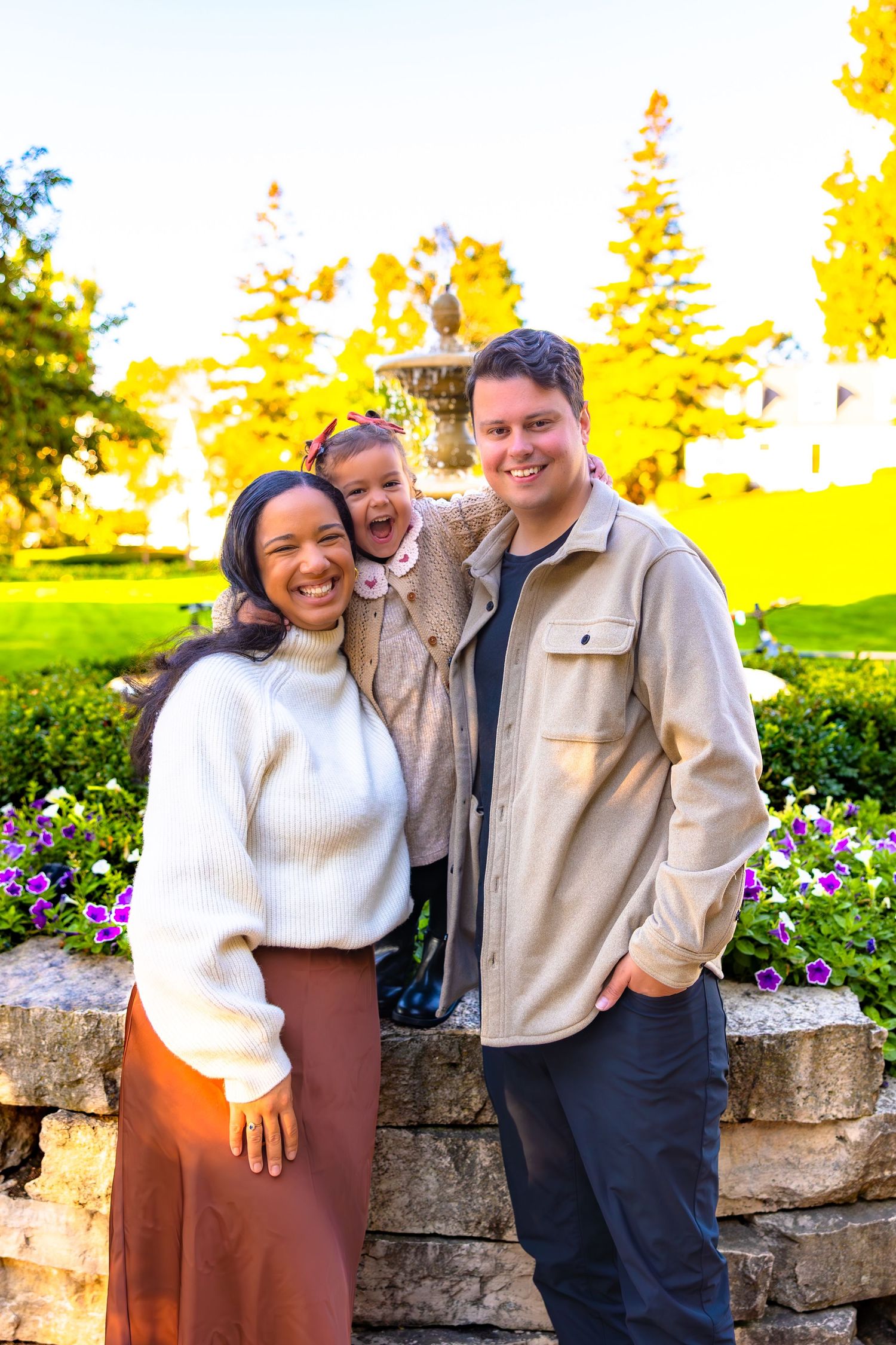 A couple poses together in front of a stone wall and garden with vibrant yellow trees in the background.