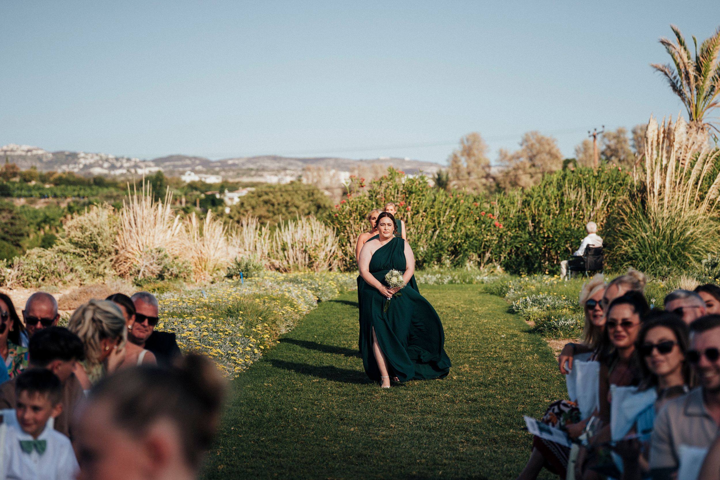 Panoramic view of an outdoor wedding ceremony with guests seated on a grassy hillside overlooking mountains at sunset.