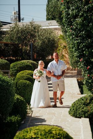 A wedding couple walks down a stone path lined with manicured hedges in a sunny garden setting.