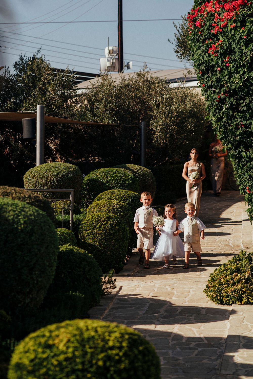 Children and adults walk along a stone path in a garden with red flowers and manicured shrubs in the background.