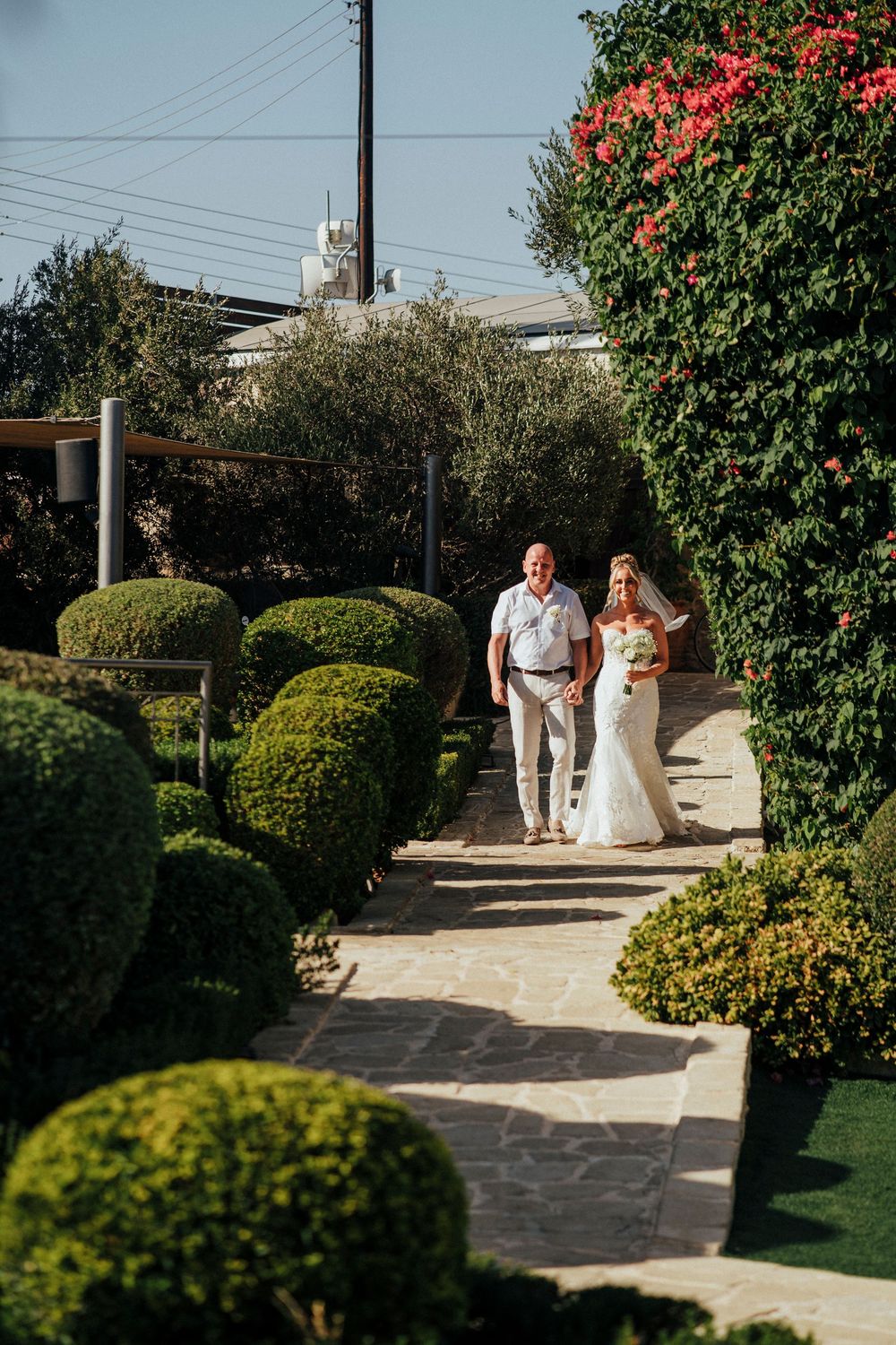 A couple walks down a garden path lined with manicured topiary bushes and flowering trees on a sunny day.