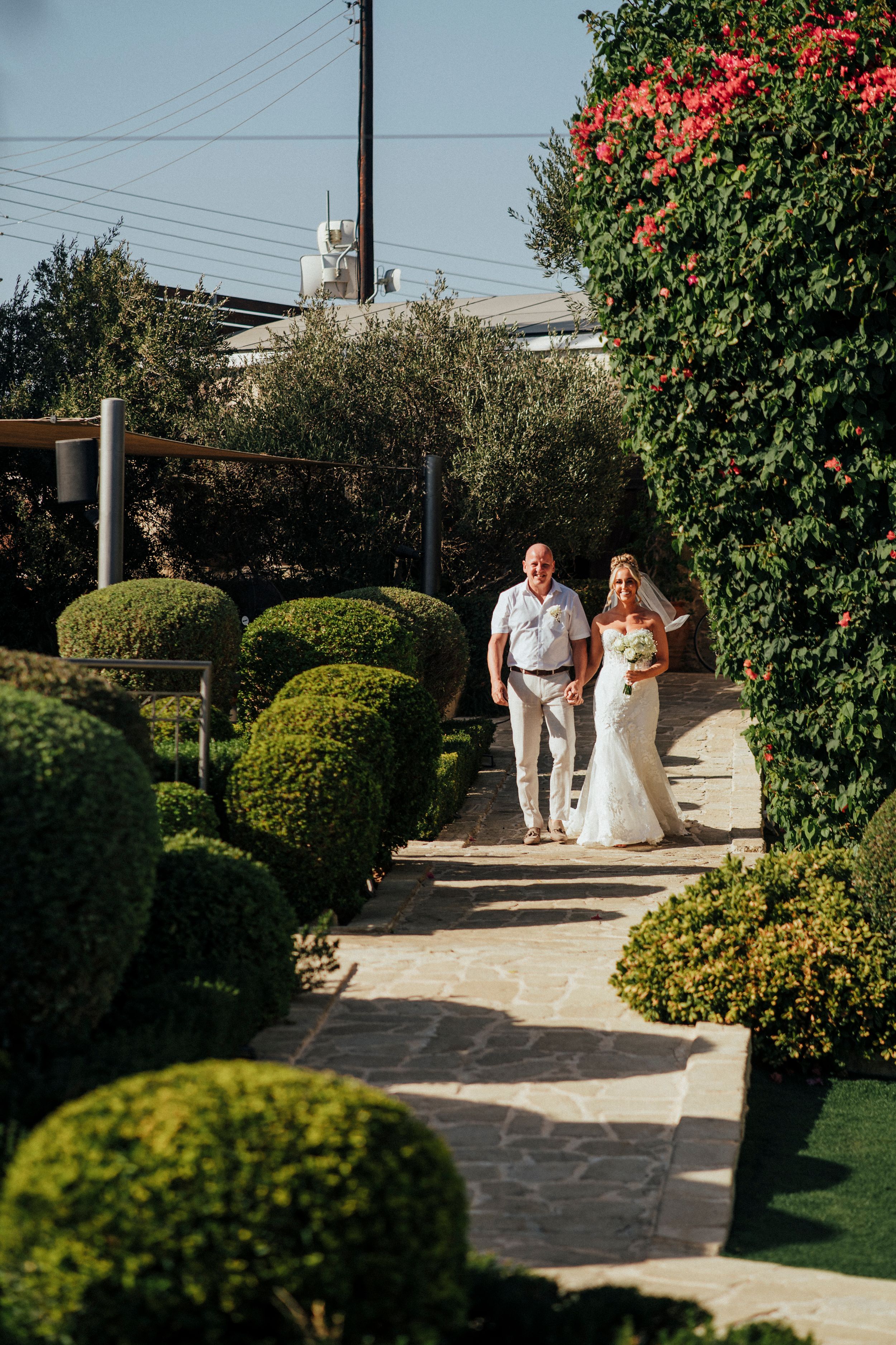 A couple walks down a garden path lined with manicured topiary bushes and flowering trees on a sunny day.