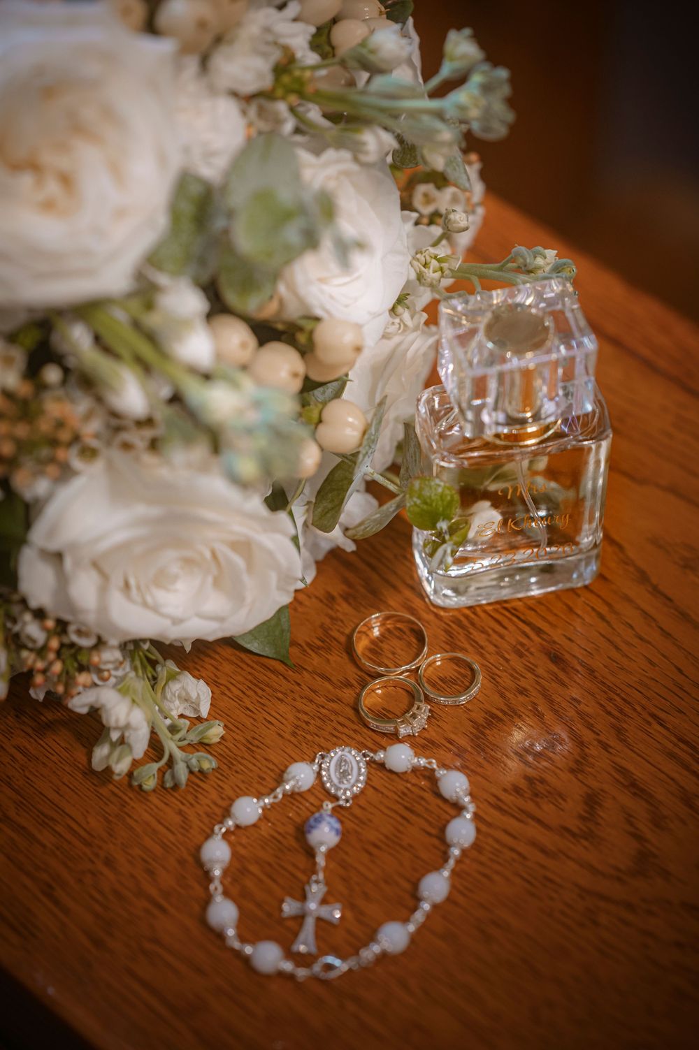 Wedding flat lay with white roses bouquet, gold rings, rosary bracelet, and crystal perfume bottle on wooden table.
