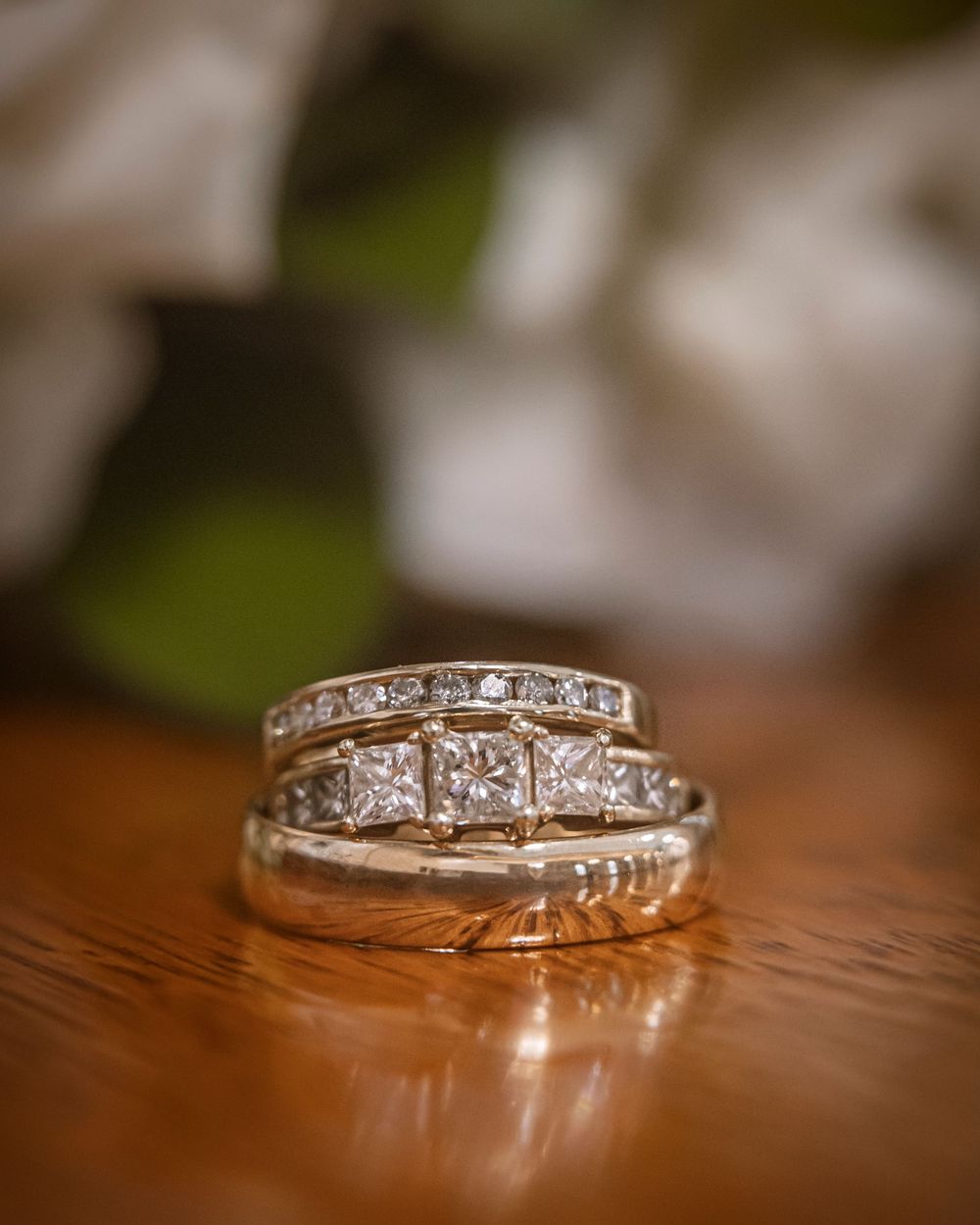 Three diamond wedding rings stacked on a wooden surface with white roses in the soft-focus background.