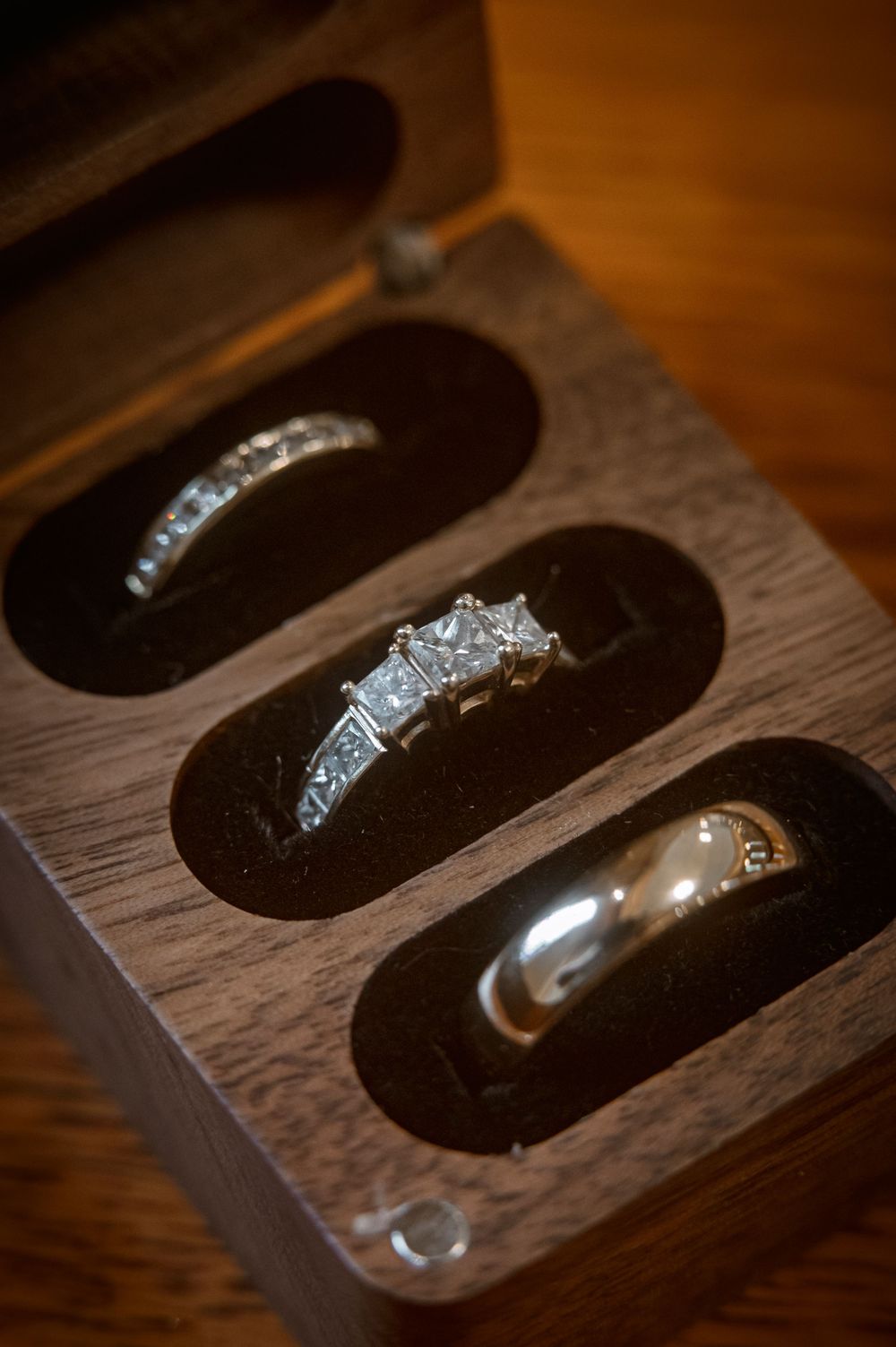 Three wedding rings displayed in a wooden jewelry box with black velvet slots, including diamond engagement ring.