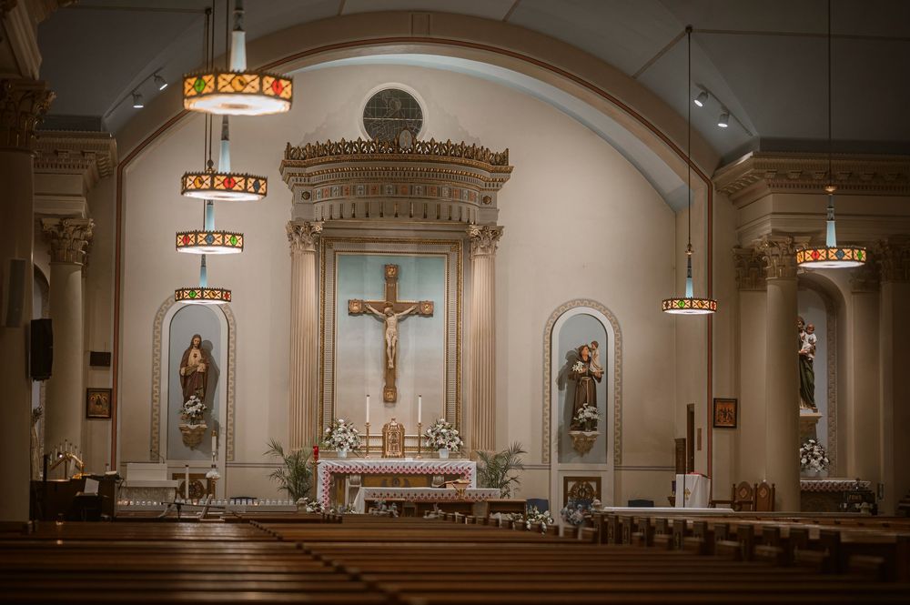 Catholic church interior with ornate altar, crucifix, religious statues, stained glass, and wooden pews in a grand sanctuary.