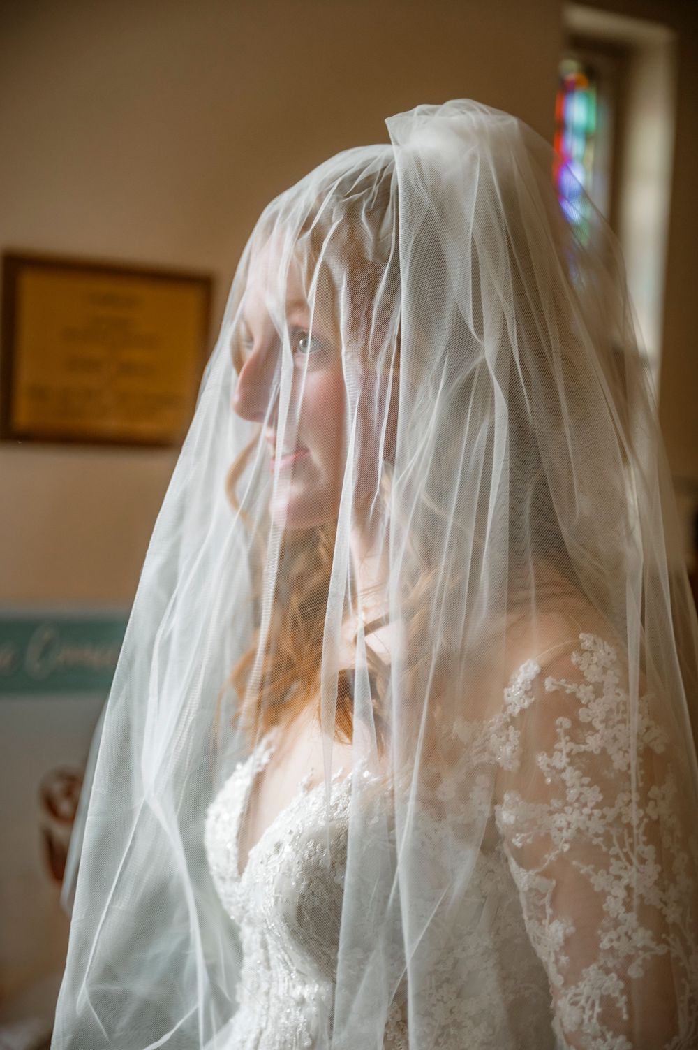 Bride wearing a lace wedding dress and sheer veil in a softly lit church, smiling gently on her wedding day.