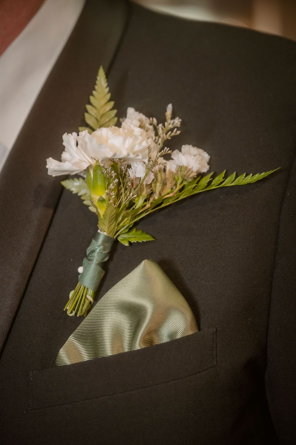 White carnation boutonniere with fern leaves and sage green pocket square on a dark suit lapel.