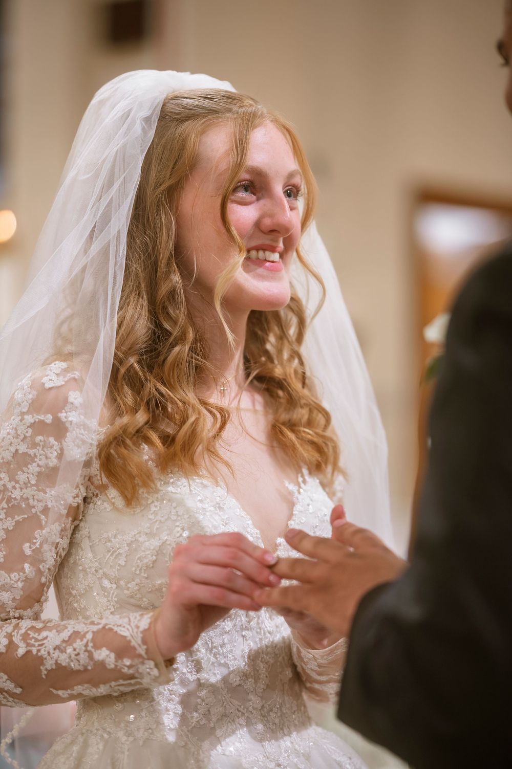 Smiling bride in lace wedding gown and veil exchanging rings at indoor ceremony.