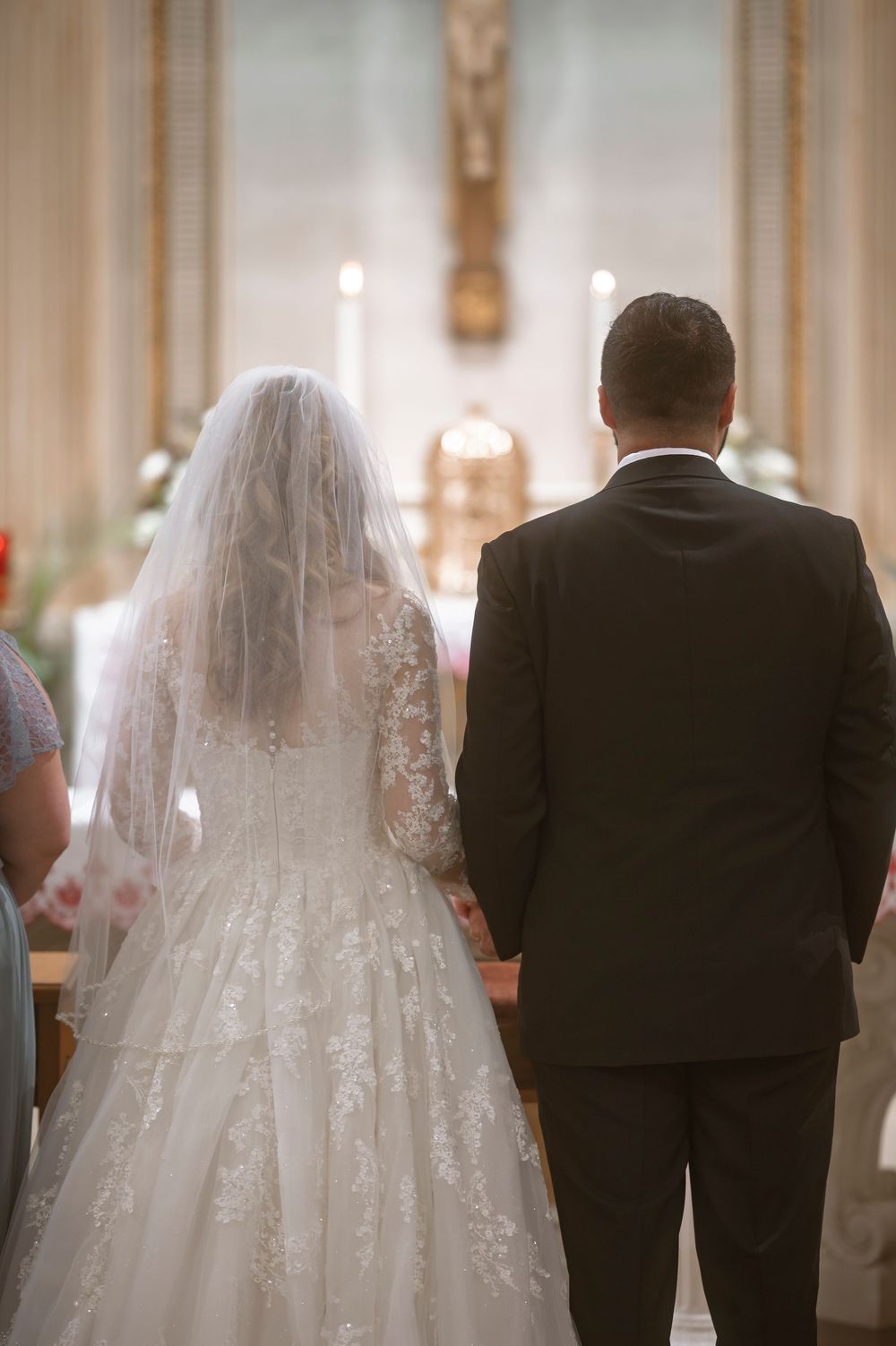 Bride in lace gown and veil stands with groom in black suit at church altar during wedding ceremony.