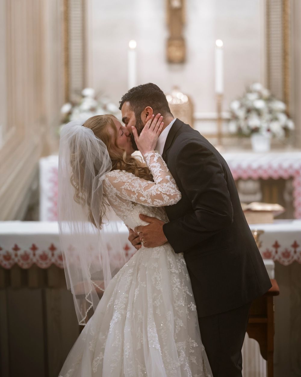 Bride and groom share first kiss at church altar, bride in lace gown with veil, groom in dark suit.