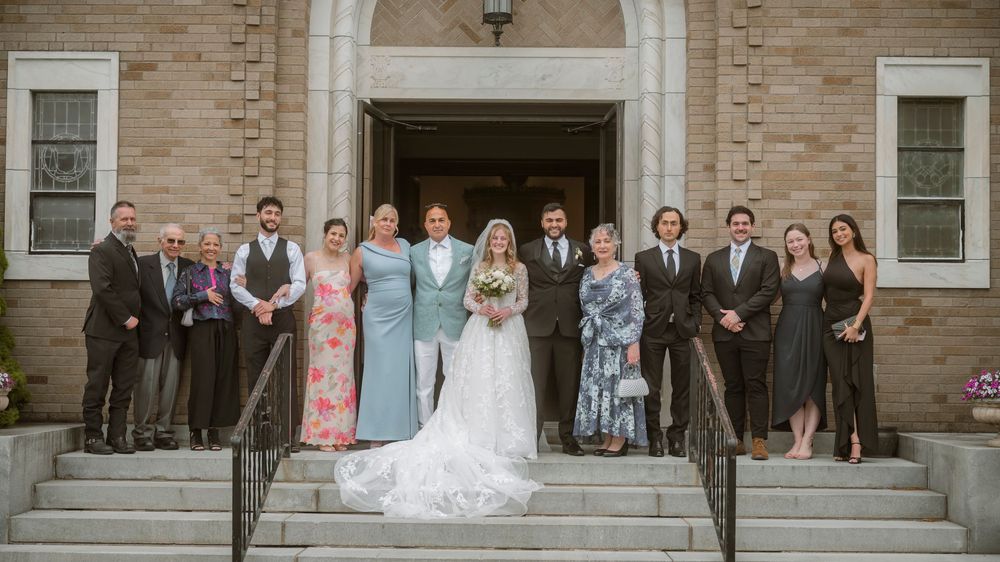 Wedding party poses on stone church steps, bride in white gown with groom and guests in formal attire.