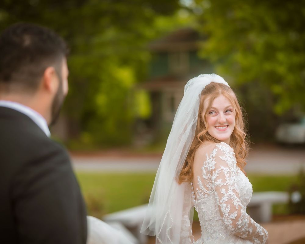 Smiling bride in lace wedding dress and veil looks back over shoulder at groom during outdoor wedding ceremony.