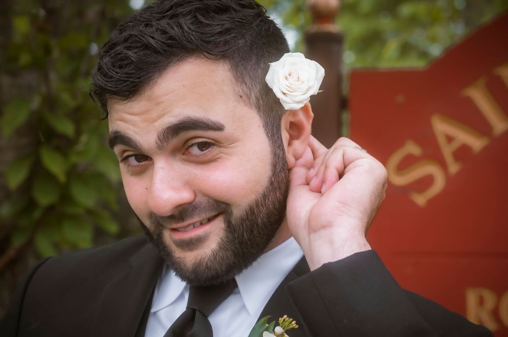 Smiling bearded groom in suit holding white rose boutonniere near his ear at outdoor wedding.