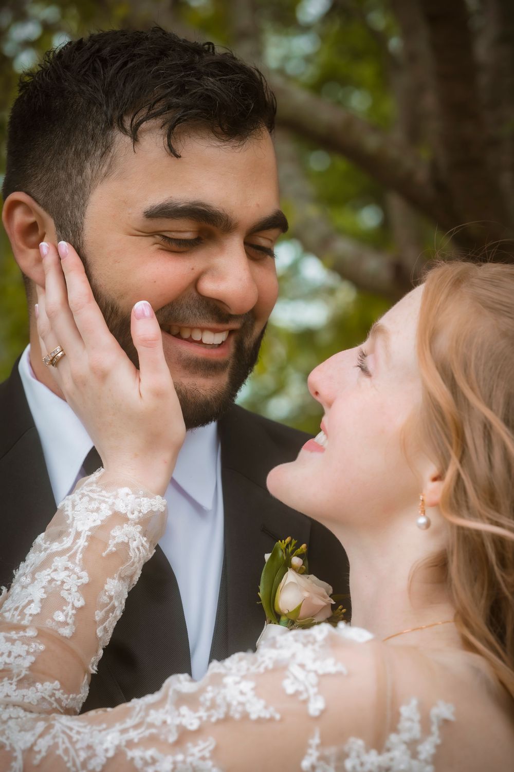 Happy bride and groom share a romantic moment, with bride in lace dress touching groom's face outdoors.