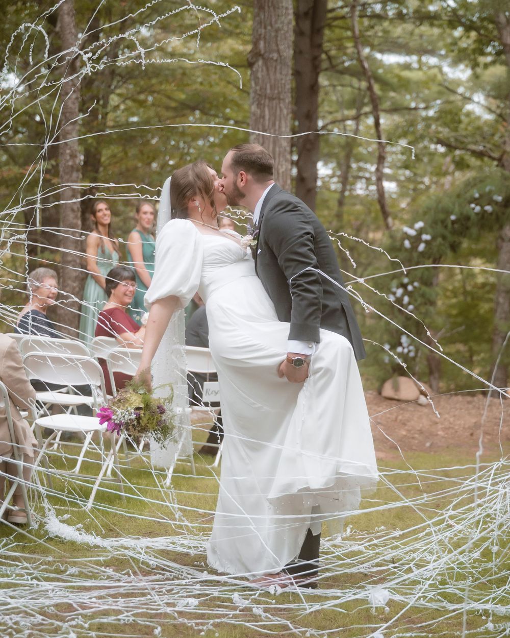 A couple laughs playfully during what appears to be an outdoor photoshoot among pine trees.