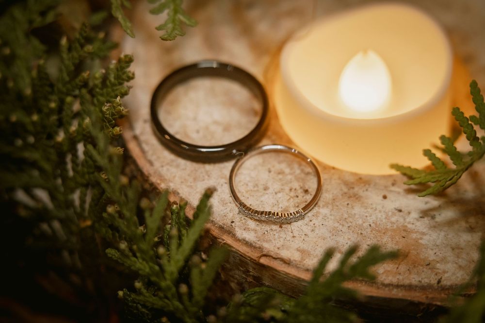Two wedding rings resting on a wooden slice beside a glowing candle surrounded by green foliage.