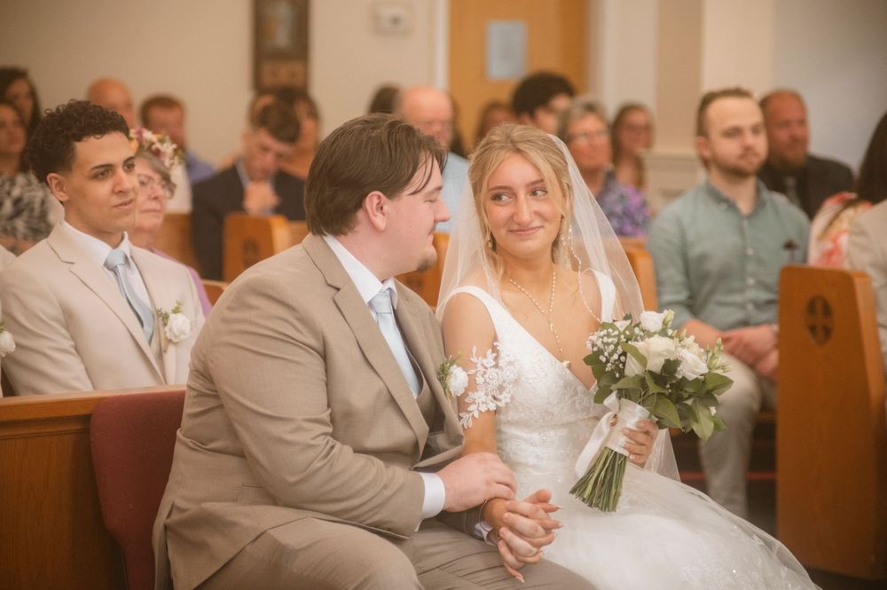 A bride and groom sit together in a church pew during their wedding ceremony while guests look on from behind.