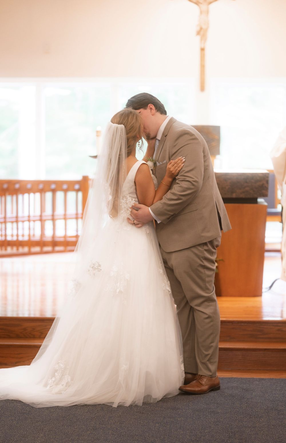 A romantic wedding moment as a couple in a tan suit and white gown embrace during their ceremony in a church.