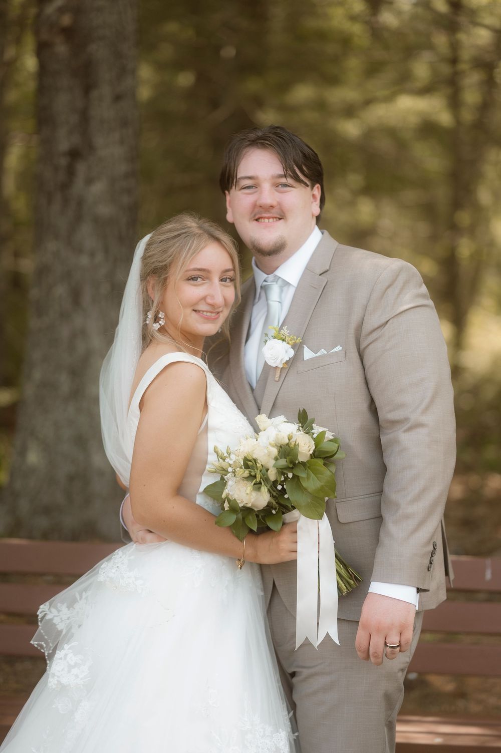 A bride and groom share intimate wedding portraits outdoors in a wooded setting with natural lighting.