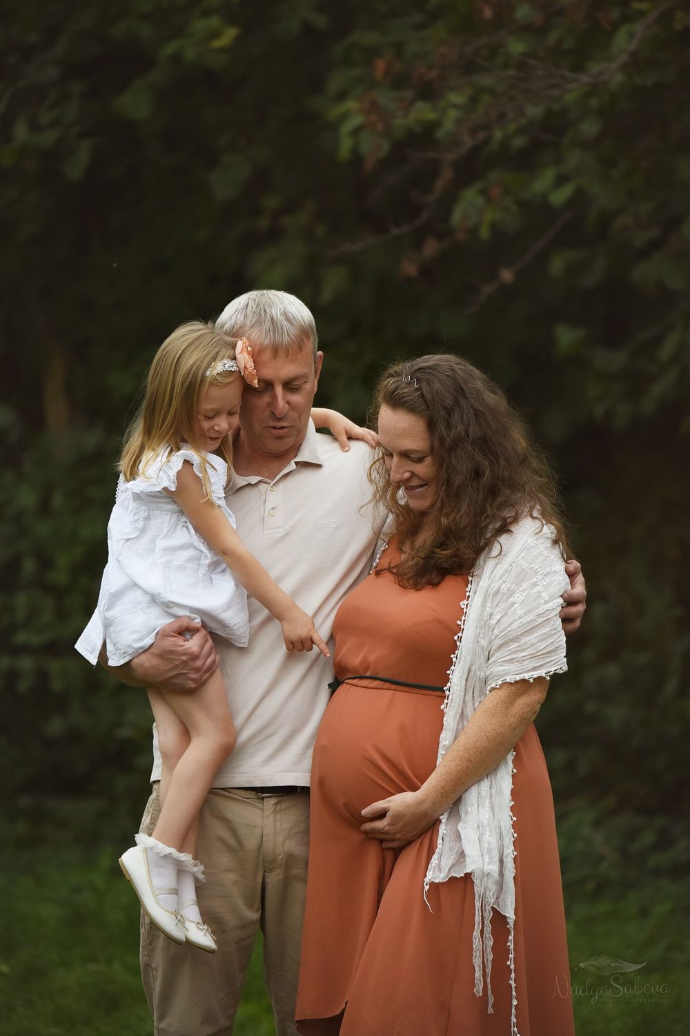 A family poses outdoors in a garden setting, with a small child in a white dress and an expectant mother in a rust-colored dress.