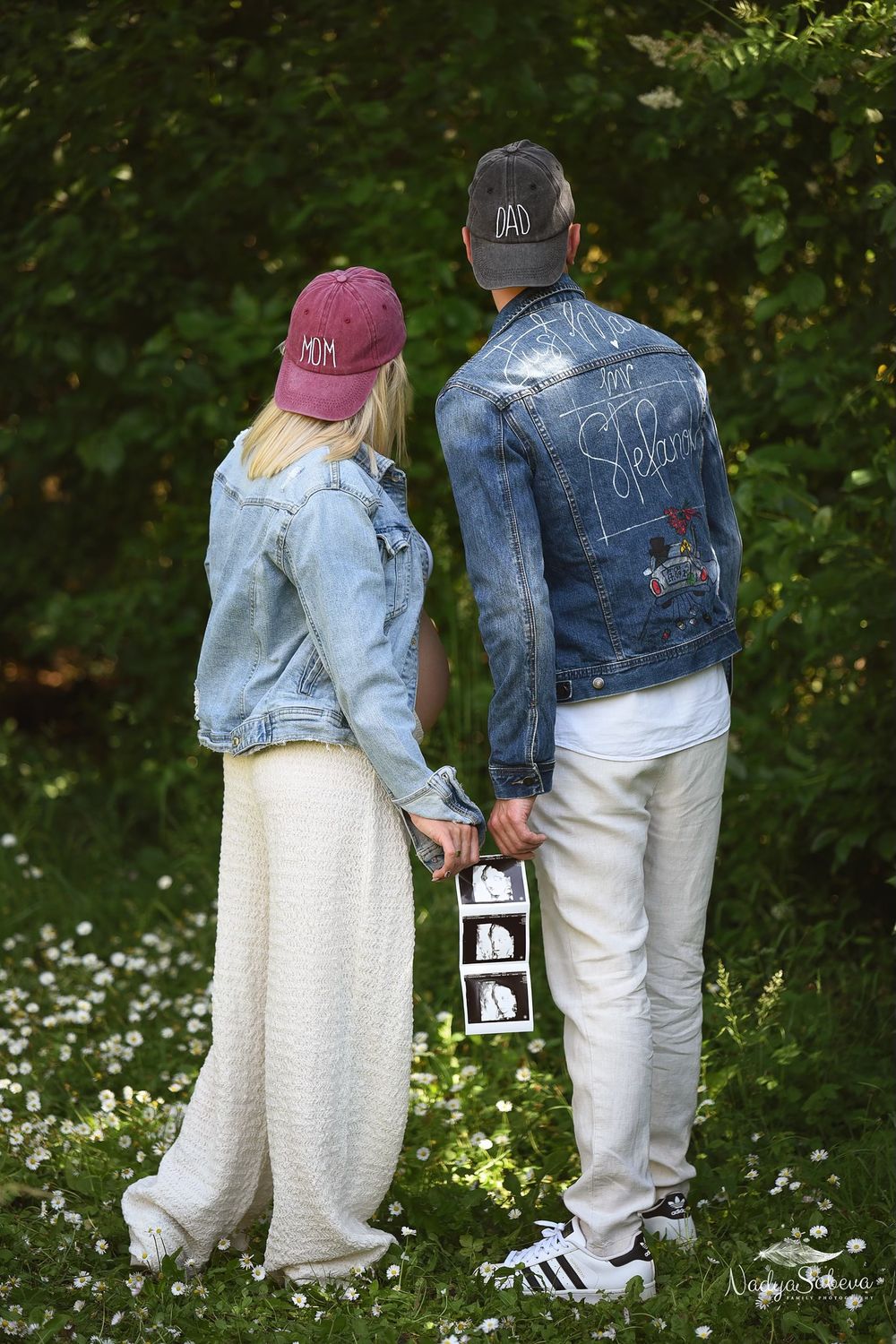 Couple in matching denim jackets and hats holding ultrasound photos in a garden with blooming flowers.