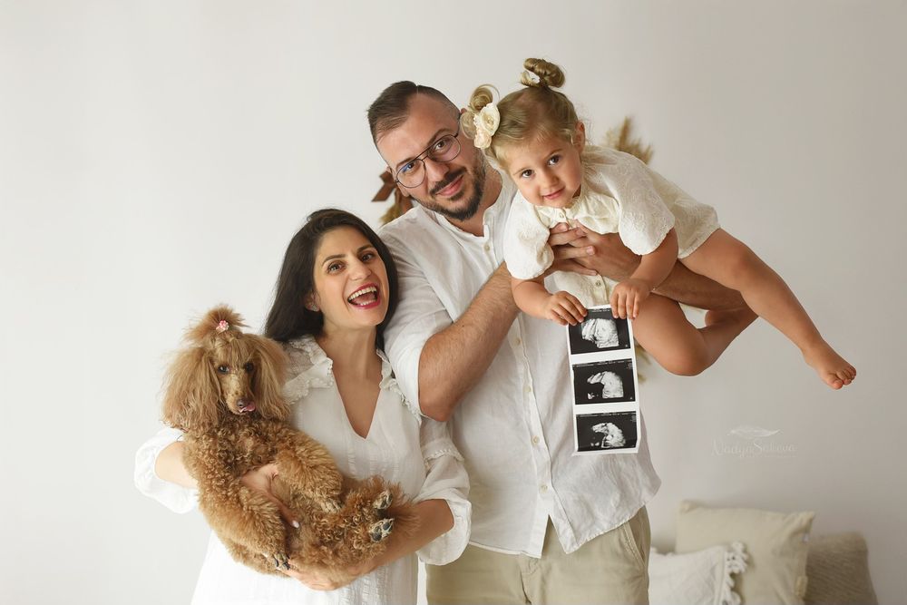 Happy family in white clothing holds teddy bear and ultrasound photo while celebrating pregnancy news against neutral background.