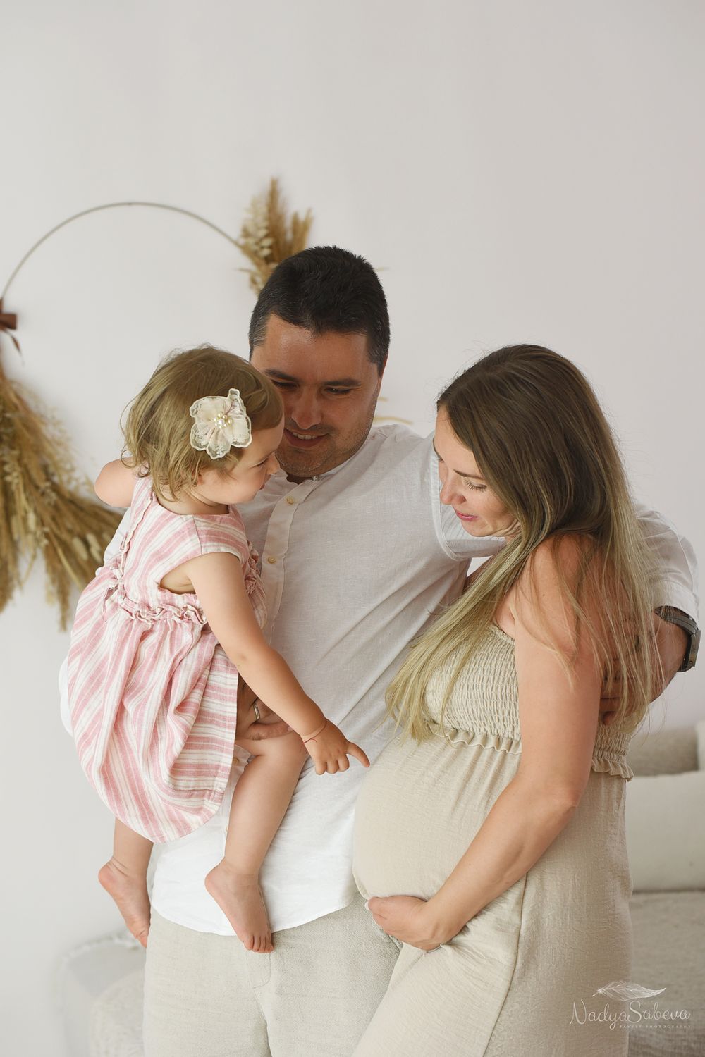 Expecting parents share a tender moment as their toddler wears a pink dress, while pampas grass decorates the backdrop.